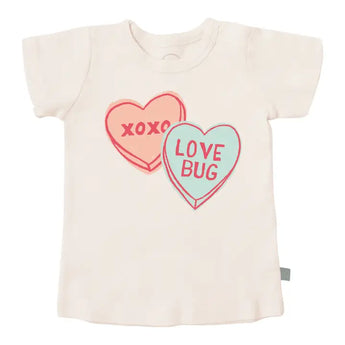 The white Organic Tee, Love Bug by Finn and Emma is a charming toddler t-shirt made from organic cotton. It features heart-shaped graphics—one pink with XOXO and the other light blue with LOVE BUG, both outlined in red using eco-friendly dyes.