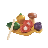 The Assorted Vegetables Set by PlanToys features a variety of wooden toy fruits and veggies like a mushroom, orange, purple fruit, two green limes, and sliced tomatoes on a wooden board with a play knife.