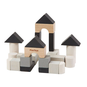 Stacked to form a castle with towers and peaked roofs, the Construction Set by PlanToys features wooden blocks in various shapes and shades of black, grey, and natural wood. It is sustainably made and displays the PlanToys label on the front block.