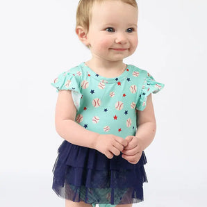 A baby smiles in a Skirt Onesie, Baseball Buddies print by Emerson and Friends, against a white background.