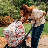 In a garden pathway surrounded by greenery and flowers, a woman with long hair wearing a white blouse and jeans leans over to check on a stroller covered with the floral-patterned Milk Snob Cover French Floral.