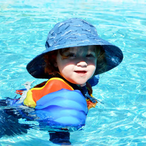 A child in a Jan and Jul Sun Hat, Shark enjoys swimming with a colorful floatation device under bright sunlight.