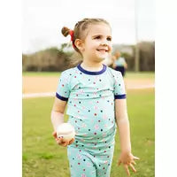 A girl in Emerson and Friends PJs, Baseball Buddies stands smiling on the field, holding a baseball and looking right.