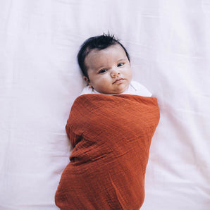 A baby rests on a soft surface, wrapped in a Rust Cotton Swaddle by Little Unicorn, gazing upward peacefully.