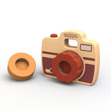 The Shutterbug Camera by Begin Again is crafted from sustainably sourced rubberwood and boasts a charming design with a red lens knob and three interchangeable side discs. Its two-toned wood features light and dark brown accents, with the brand name elegantly displayed on top.