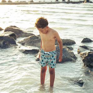 A boy in Emerson and Friends Pirates Life swim trunks stands by rocks, sunlight reflecting on the sea.