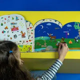 A child with long hair is playing with a Floss and Rock Stick and Play, Adventure mounted on a yellow board. The playbook features colorful animal illustrations and outdoor scenes on a blue background while the child places an adventure sticker on it.