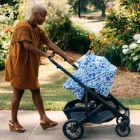 In a garden, someone in a brown dress with short hair and platform sandals pushes a stroller along the sidewalk. The stroller, elegantly shielded by the Milk Snob Cover in Disneys Stitch design, features blue and white hues against the colorful backdrop of greenery and flowers.