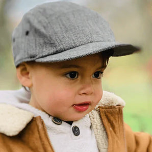 A child in a Storied Hats Lil Hat, Tuxedo, wears a brown jacket with a fluffy collar, glancing to the side outdoors.