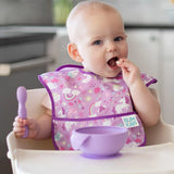 Baby in high chair, unicorn bib on, using Bumkins Silicone First Feeding Set: Lavender, purple bowl on tray.