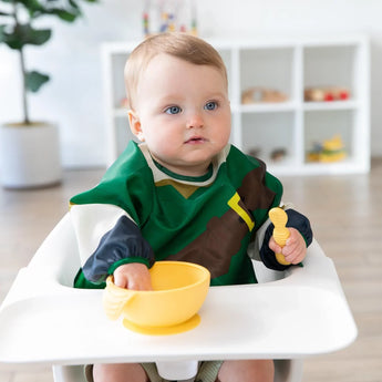 A baby in a high chair holds a yellow bowl and spoon, wearing the waterproof Zelda™ Sleeved Bib by Bumkins. Shelves and a plant behind.