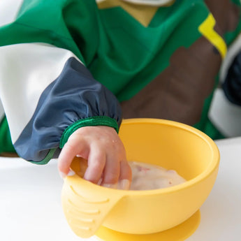 A childs hand reaches into a yellow bowl, wearing a Bumkins Zelda™ Sleeved Bib, made from waterproof, easy-clean fabric.