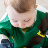 A serene baby with light hair in a green Bumkins Sleeved Bib: Zelda™, featuring brown and yellow patterns, is the focus. Highlighting their peaceful expression and long eyelashes, this moment of calm is enhanced by the bibs waterproof fabric, ensuring easy cleaning for busy parents.