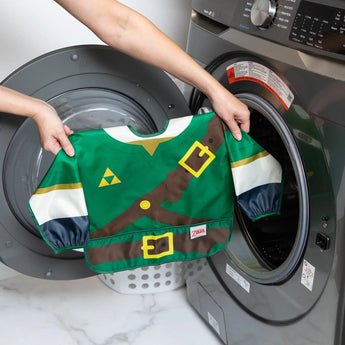 Two hands gently load a Bumkins Sleeved Bib: Zelda™ into a front-load washer for easy cleaning.