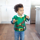 Young child wears Bumkins Zelda™ sleeved bib in green with triangles, made of waterproof fabric, standing in a kitchen.