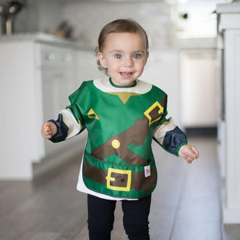 A toddler beams in the kitchen wearing a Bumkins Sleeved Bib: Zelda™, an adventure-themed, green and brown waterproof costume.