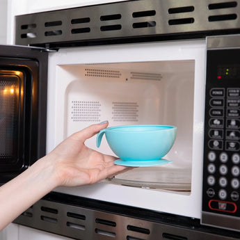 A hand places a blue bowl from Bumkins Silicone First Feeding Set into a microwave, showing its interior and right control panel. Made of food-safe silicone, the bowl includes a handle and suction base for easy self-feeding.