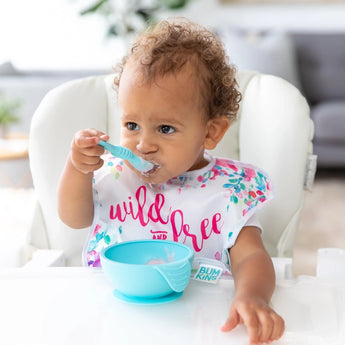 A toddler in a high chair enjoys a meal with their Bumkins silicone spoon from the Blue First Feeding Set and wild and free on their bib.