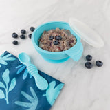 The Bumkins Silicone First Feeding Set in blue is perfect for self-feeding toddlers with its bowl, lid, and spoon on a marble surface.