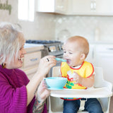 In a bright kitchen, a baby in a high chair is fed with Bumkins Silicone First Feeding Set: Blue, featuring a food-safe spoon and bowl.