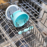 A Bumkins Silicone First Feeding Set in blue, featuring a light blue baby bowl and spoon, is placed on the top rack of an open dishwasher. The bowl is upside down next to the spoon among the metal prongs.