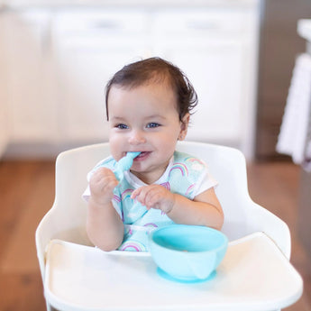 Baby in high chair with rainbow bib, smiling and holding a blue spoon from the Bumkins Silicone First Feeding Set: Blue.