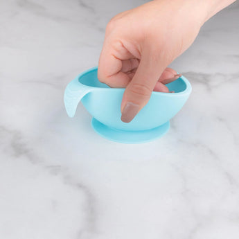 A hand holds Bumkins Silicone First Feeding Set: Blue, a food-safe silicone bowl with a suction base on a marble surface.