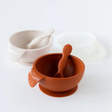 The Bumkins Silicone First Feeding Set: Clay includes two bowls with matching spoons in rust and cream, each with a suction base and lid.