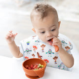 Baby in a bib with cartoons uses Bumkins Silicone First Feeding Set: Clay spoon to enjoy fruit pieces from an orange bowl.