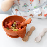 A toddler grabs the Bumkins Silicone First Feeding Set: Clay, with fruit pieces and two beige baby spoons on a white table.