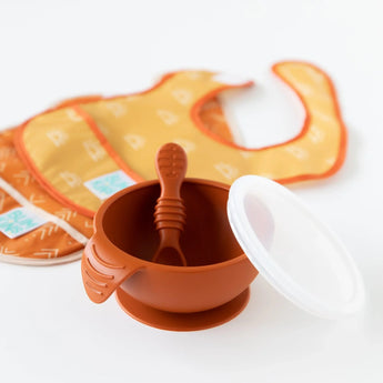 Bumkins Silicone First Feeding Set: Clay includes a brown suction bowl, baby spoon with a white lid, and an orange geometric-patterned bib.