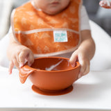 A baby in an orange bib uses the Bumkins Silicone First Feeding Set: Clay, gripping the handle with determination to self-feed.