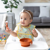 A baby in a high chair, with a white bow, mastering self-feeding using Bumkins Silicone First Feeding Set: Clay and a baby spoon.