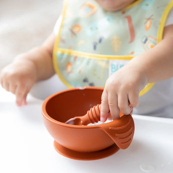 A baby in a bib grips Bumkins Silicone First Feeding Set: Clay, with a tiny spoon nestled in the brown bowl on the tray.