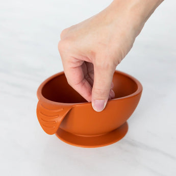 Holding the Bumkins Silicone First Feeding Set: Clay, a small orange bowl with suction, perfect for self-feeding with a baby spoon.