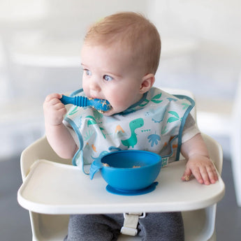 Baby in high chair using Bumkins Gumdrop spoon, wearing dinosaur bib. Blue bowl on tray nearby.