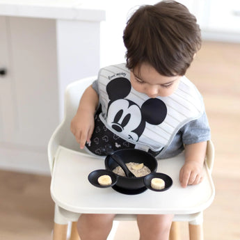 A toddler enjoys breakfast in a high chair with Bumkins Mickey Mouse Classic Black Silicone First Feeding Set, featuring a Mickey-shaped bowl.