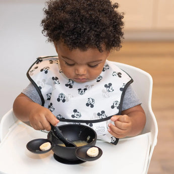 A toddler in a high chair enjoys bananas with a Bumkins Mickey Mouse Classic Black silicone spoon from the feeding set.