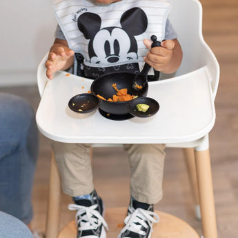 A child in a high chair enjoys mashed food with a spoon, wearing a Bumkins Disney bib, with the Mickey Mouse Classic Black feeding set.