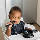 A toddler in a high chair, wearing a bib from the Disney Black + White Collection, uses Bumkins Mickey Mouse Classic Black feeding set.