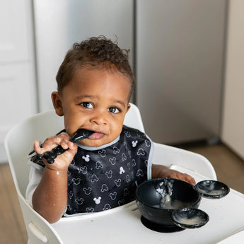 A toddler in a high chair, wearing a bib from the Disney Black + White Collection, uses Bumkins Mickey Mouse Classic Black feeding set.