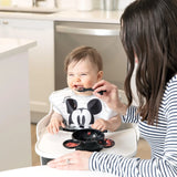 Adult feeds grinning baby in striped tops; baby clutches a utensil with a Bumkins Mickey Mouse Black Bib from Disneys Collection.