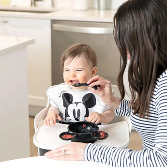 Adult feeds grinning baby in striped tops; baby clutches a utensil with a Bumkins Mickey Mouse Black Bib from Disneys Collection.