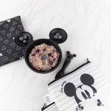 Mickey Mouse-shaped bowl with oatmeal, blueberries, spoon, & Bumkins Disney Black + White Collection pouches on a white marble surface.