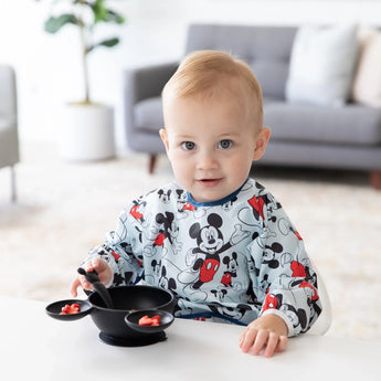 A baby in a Mickey Mouse outfit uses the Bumkins Silicone First Feeding Set: Mickey Mouse Classic Black at a bright, modern table.