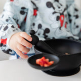 A child in a Mickey Mouse shirt holds a Bumkins Silicone First Feeding Set utensil over the matching bowl filled with small food pieces.