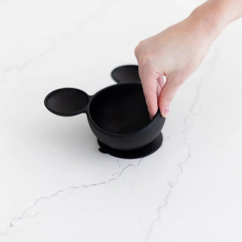 A hand holds the Bumkins Silicone Feeding Set: Mickey Mouse Classic Black, featuring mouse ear handles, on a white marble surface.