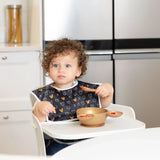 A toddler with curly hair and Disney Minnie Mouse ears uses Bumkins Silicone First Feeding Set: Minnie Mouse Gold in a modern kitchen.