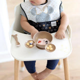 A baby in a high chair explores the Bumkins Minnie Mouse Gold Silicone First Feeding Set with yogurt, berries, and snacks nearby.