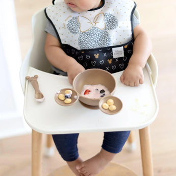 A baby in a high chair explores the Bumkins Minnie Mouse Gold Silicone First Feeding Set with yogurt, berries, and snacks nearby.